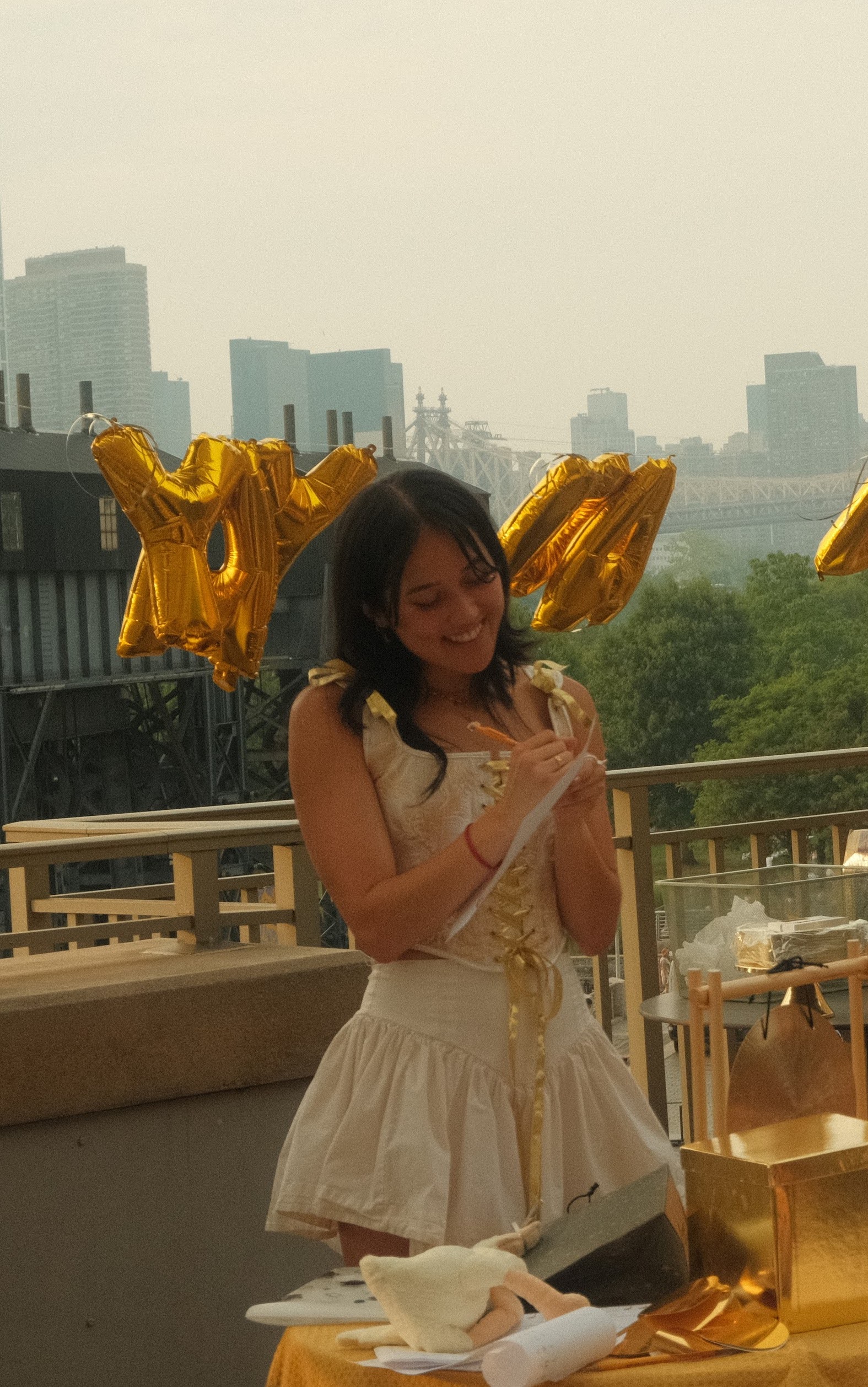 Heidi in gold top and white skort standing and writing on a piece of paper in front of gold balloons.