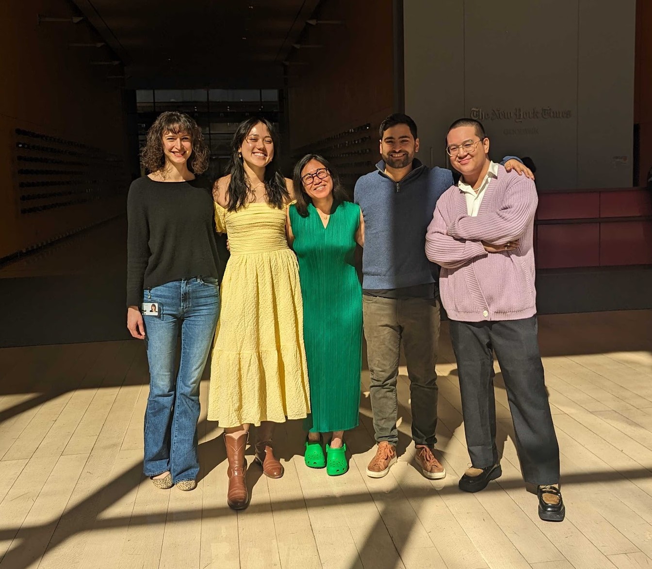 In order from left to right: Zoe Bell, Heidi Erwin, Wyna Liu, Joel Fagliano, and Robert Vinluan stand in the lobby of The New York Times building. Zoe is wearing black, Heidi yellow, Wyna green, Joel blue, and Robert purple, repping the colors of the NYT Connections game.